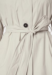 Trench-coat beige avec une ceinture nouée, une fermeture à boutons et un tissu texturé. Comprend des poches latérales et une finition lisse.