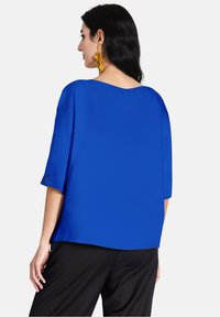 Top blu loose-fitting con maniche corte e scollatura ampia. Tessuto liscio con orlo dritto, indossato con pantaloni neri.