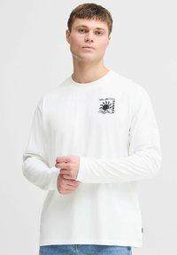 Camiseta de manga larga blanca hecha de algodón con un gráfico negro que presenta el texto "VERANO ILIMITADO" y un ícono de sol en el lado izquierdo del pecho.