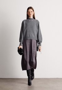 Γκρι πλεκτή μπλούζα με μπαλόνι μανίκια, σε συνδυασμό με ένα σκούρο μωβ midi φόρεμα που διαθέτει πλάγιες κορδέλες. Το μοντέλο κρατά μια μαύρη τσάντα.