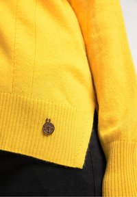 Žlutý pletený svetr s proužkovaným lemem a rukávy. Obsahuje malý bronzový přívěsek ve tvaru kotvy poblíž lemu. Měkká textura s rovnoměrným šitím.