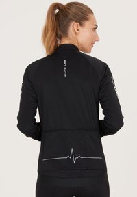Zwarte sportjack met een hoge kraag, gedrukt logo op de achterkant en een grafische hartslaglijn op de onderste achterkant. Geweven, aansluitend ontwerp.
