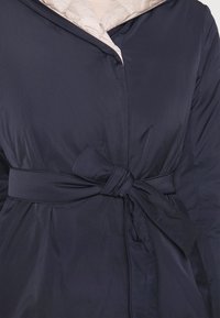 Manteau bleu marine ceinturé avec un nœud à la taille et une doublure intérieure beige clair contrastante, porté sur un haut clair.