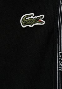 Πράσινο κεντημένο λογότυπο κροκόδειλου με λευκό περίγραμμα σε μαύρο ύφασμα δίπλα σε μαύρη λωρίδα με λευκό κείμενο "LACOSTE" και ρίγες.