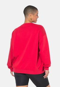 Sudadera roja de ajuste holgado, con puños y dobladillo acanalados. Textura suave, cuello redondo y diseño de hombros caídos. Llevada con pantalones cortos negros.