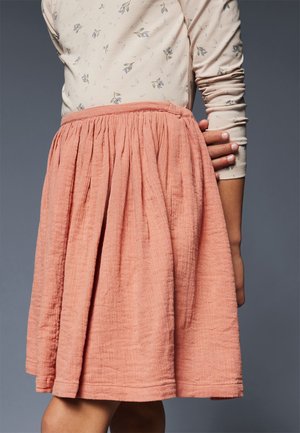 Ragazza che indossa una gonna a pieghe color pesco e una camicia beige a maniche lunghe con piccoli motivi floreali, in piedi contro uno sfondo grigio.