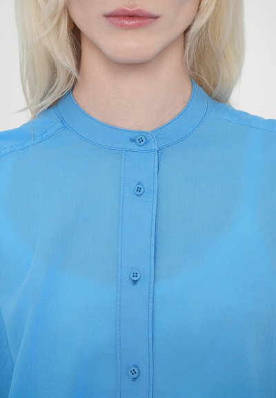 Camicia azzurra chiara con colletto tondo, dotata di quattro bottoni sulla parte anteriore e realizzata in un tessuto morbido e leggero.