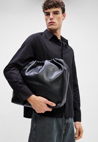 Mustast nahast nööriga kott, millel on sile tekstuur, pehme ja ümar kuju, hoituna musta mustriga särgi ja teksade vastu.