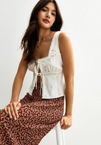 Witte crop top met bloemenborduring, verstelbare voorknoop en kantdetails. Gecombineerd met een lange bordeauxrode bloemhoudende rok.