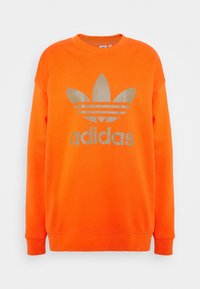 Sudadera naranja hecha de una mezcla de algodón, con un gran logo metálico dorado de Adidas y el icónico diseño de las tres franjas en el frente.