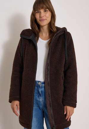 Femme portant une longue veste polaire marron foncé avec capuche, chemise blanche et jean bleu, debout devant un fond clair uni.