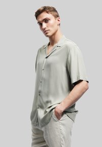 Lichtgroen, kort mouwen shirt gemaakt van gladde stof, met een knoopsluiting en zachte kraag, gedragen over lichtgekleurde broeken.