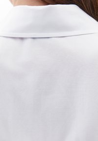 Hvid skjorte med en glat, tekstureret stof; har en klassisk krave og et rent, minimalistisk design. Ingen synlige mønstre eller dekorationer.