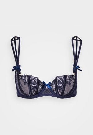 Námořnická modrá krajková podprsenka s průsvitnými košíčky, dekorativními mašlemi na ramínkách a vlnkovými okraji. Ramínka mají geometrický design.