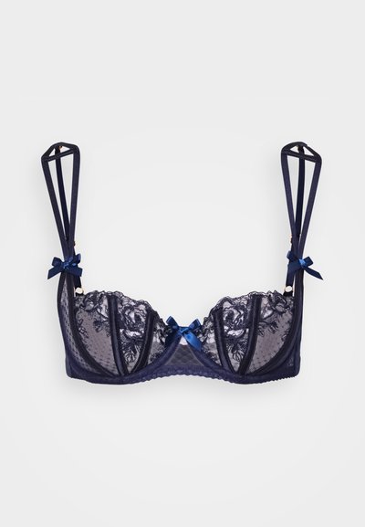 Jūras zilas mežģīņu krūšturis ar caurspīdīgām kausām, dekoratīvām lentītēm uz siksnām un viļņotām malām. Siksnām ir ģeometriska izgriezuma dizains.