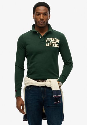 Mand iført en mørkegrøn langærmet polo med teksten "Superdry Athletics", mørke jeans og en hvid sweater bundet om taljen.