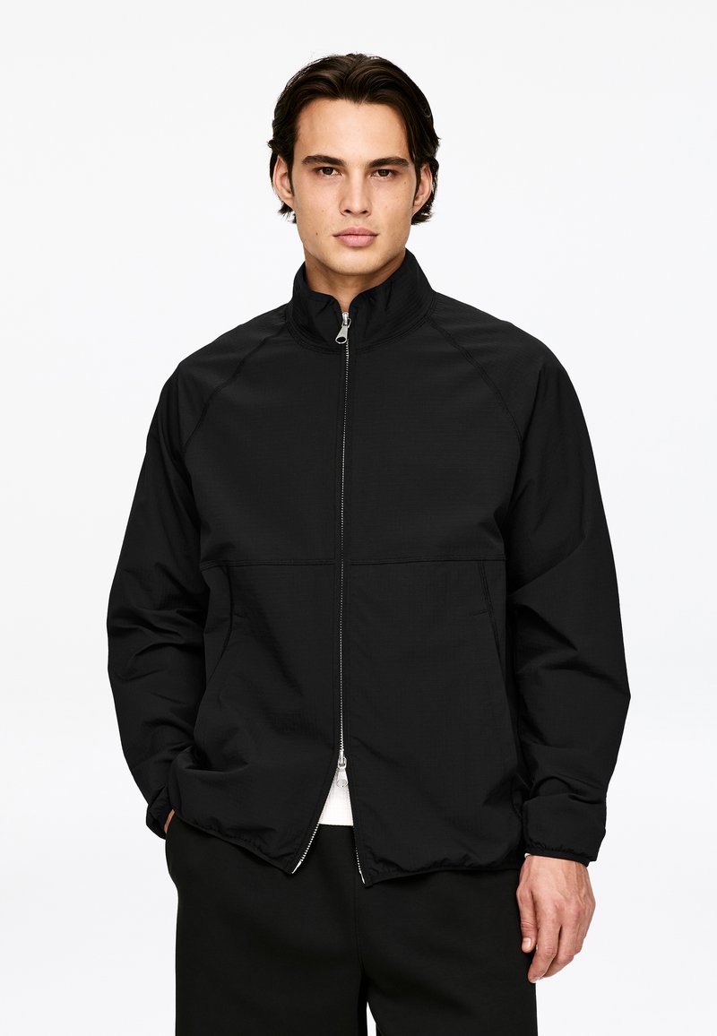 Young man with dark hair wearing a black zip-up jacket and black pants, standing against a plain white background.