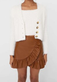 Weißer flauschiger, kurzer Cardigan mit großen Knöpfen über einem passenden flauschigen Top, kombiniert mit einem braunen, rüschierten Minirock aus Kunstleder.