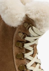 Braune Wildlederstiefel mit einem flauschigen cremefarbenen Fellkragen, ausgestattet mit metallischen Ösen und hellbeigen Schnürsenkeln zur Schließung. Strukturierte Oberflächenbeschreibung sichtbar.