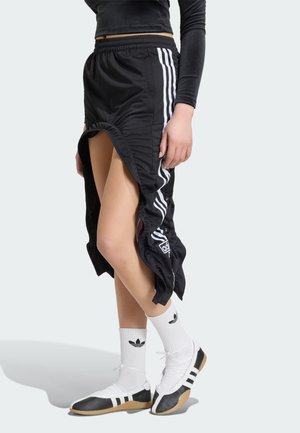 Osoba nosząca czarne spodnie dresowe Adidas z otwartymi bocznymi zatrzaskami odsłaniającymi nogi, w połączeniu z białymi skarpetkami Adidas i czarno-białymi butami.