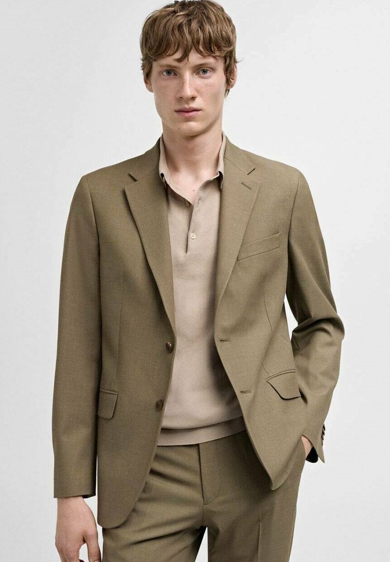 Hellbrauner Anzug mit schlanker Passform, einreihigem Sakko und passenden Hosen. Kombiniert mit einem beigen Poloshirt. Glatte Stoffstruktur.