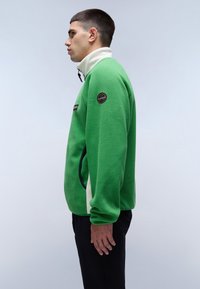 Groene fleecetruien met hoge kraag, witte accenten, zijzakken en een rond logo-patch op de linkermouw. Zachte textuur, casual pasvorm.
