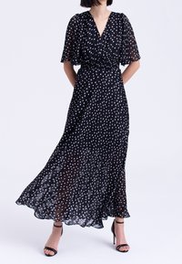 Svart maximalång klänning med vita prickar, korta volangärmar och smockdetalj vid midjan. Tillverkad av lätt, genomskinlig tyg som flödar.