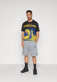 Μαύρο και κίτρινο γραφικό μπλουζάκι με αθλητικό λογότυπο, συνδυασμένο με γκρι σορτς. Μαύρα παπούτσια και κάλτσες ολοκληρώνουν την αθλητική εμφάνιση.