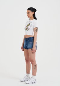 Witte crop top met bloemen- en bijenprint, gecombineerd met blauwe spijkershorts. Witte sneakers met grijze accenten en zichtbare texturen.