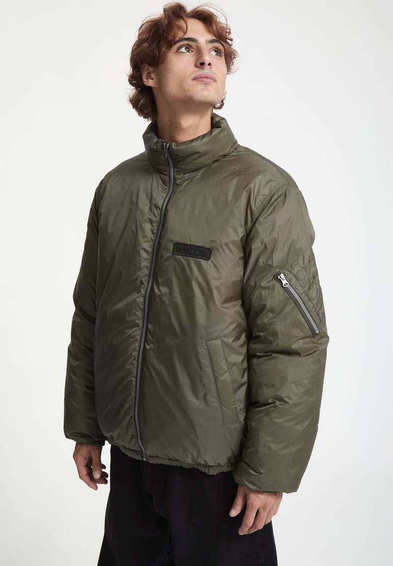 Chaqueta acolchada verde oliva con cuello alto, cierre de cremallera y acentos acolchados en los hombros. Presenta un parche con el logo en el frente y bolsillos laterales.