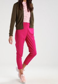 Olivgrön quiltad bomberjacka över en ljusrosa skjorta med krage, parat med klara rosa tapered byxor och pastellrosa sneakers med accenter.