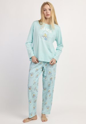 Joven de pie descalza, vestida con un conjunto de camiseta de manga larga y pantalones azul claro con un estampado de dibujos animados de Alicia en el País de las Maravillas.