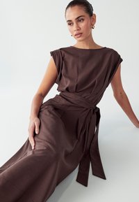 Hnědé bezramínkové šaty s volným střihem, širokým lodním výstřihem a detailem s vázáním v pase, vyrobené z hladkého, texturovaného materiálu.