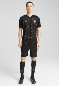 Camiseta de fútbol negra con patrón agrietado naranja, de manga corta y cuello en V, combinada con pantalones cortos y calcetines negros, con acentos y logo en naranja.