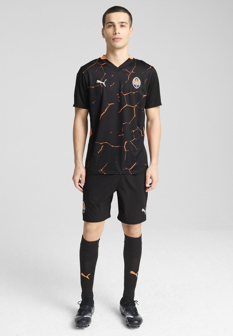 Camiseta de fútbol negra con patrón agrietado naranja, de manga corta y cuello en V, combinada con pantalones cortos y calcetines negros, con acentos y logo en naranja.