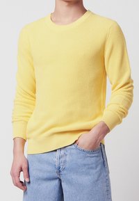 Gelber Strickpullover mit rundem Halsausschnitt, gerippten Bündchen und einem strukturierten Muster, kombiniert mit hellblauen Jeans.