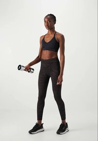 Žena u crnom sportskom grudnjaku i leggingama s uzorkom, drži bučicu jednom rukom, stojeći na jednostavnoj svijetloj pozadini.
