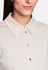 Light beige blouse featuring a structured collar, button front with decorative buttons, and soft, smooth fabric.