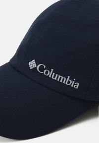 Granatowa czapka baseballowa wykonana z miękkiego materiału, z zakrzywionym daszkiem i srebrnym logo "Columbia" z przodu. Widoczne detale szwów.