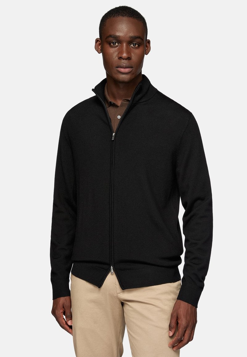 Black knitted zip-up sweater with a high collar, ribbed texture, and zipper hardware, worn over a brown collared shirt, paired with beige pants.