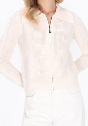 Femme portant un pull en maille crème zippé avec un large col et un pantalon blanc, les mains reposant dans le dos.