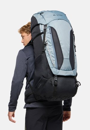 Jeune homme portant un grand sac à dos de randonnée bleu et noir avec plusieurs sangles et compartiments, vu de profil et de dos.