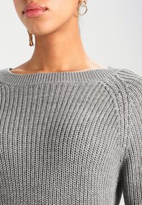 Närbild av en person som bär en grå ribbad stickad tröja och guldiga hoopsörhängen mot en ljusgrå bakgrund.