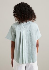 Camicia a maniche corte a righe blu chiaro e bianche con dettaglio a pieghe sulla schiena e orlo arrotondato, realizzata in un tessuto morbido.