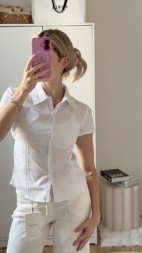 Camisa branca de manga curta, com um design ajustado, apresentando costuras verticais subtis e um colarinho clássico, combinada com calças bordadas claras.