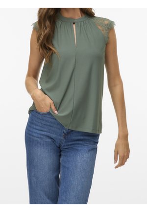 Femme portant un haut vert sauge sans manches avec des épaules en dentelle et un décolleté en forme de serrure, associé à un jean bleu en denim, main dans la poche.