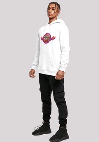 Sudadera blanca con un logo circular de "Wonder Woman", bolsillo frontal y capucha con cordón, combinada con pantalones cargo negros y botas negras.