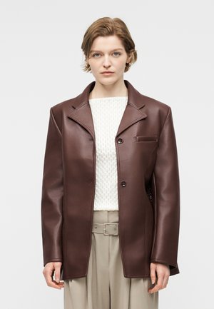 Γυναίκα που φοράει καφέ δερμάτινο σακάκι πάνω από λευκό πλεκτό πουλόβερ και μπεζ παντελόνια με ζώνη, στέκεται μπροστά σε απλό φωτεινό φόντο.