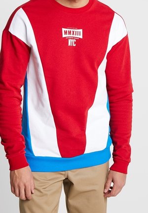 Mężczyzna w bluzie w kolorach czerwonym, białym i niebieskim z napisem "MMXIII NYC" na piersi oraz beżowych spodniach.