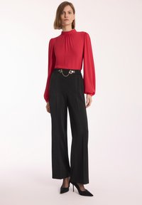 Czerwona bluzka z długimi rękawami i wysokim kołnierzem, zestawiona z czarnymi spodniami z szerokimi nogawkami, które mają łańcuszek na pasku. Czarne szpilki w szpic.